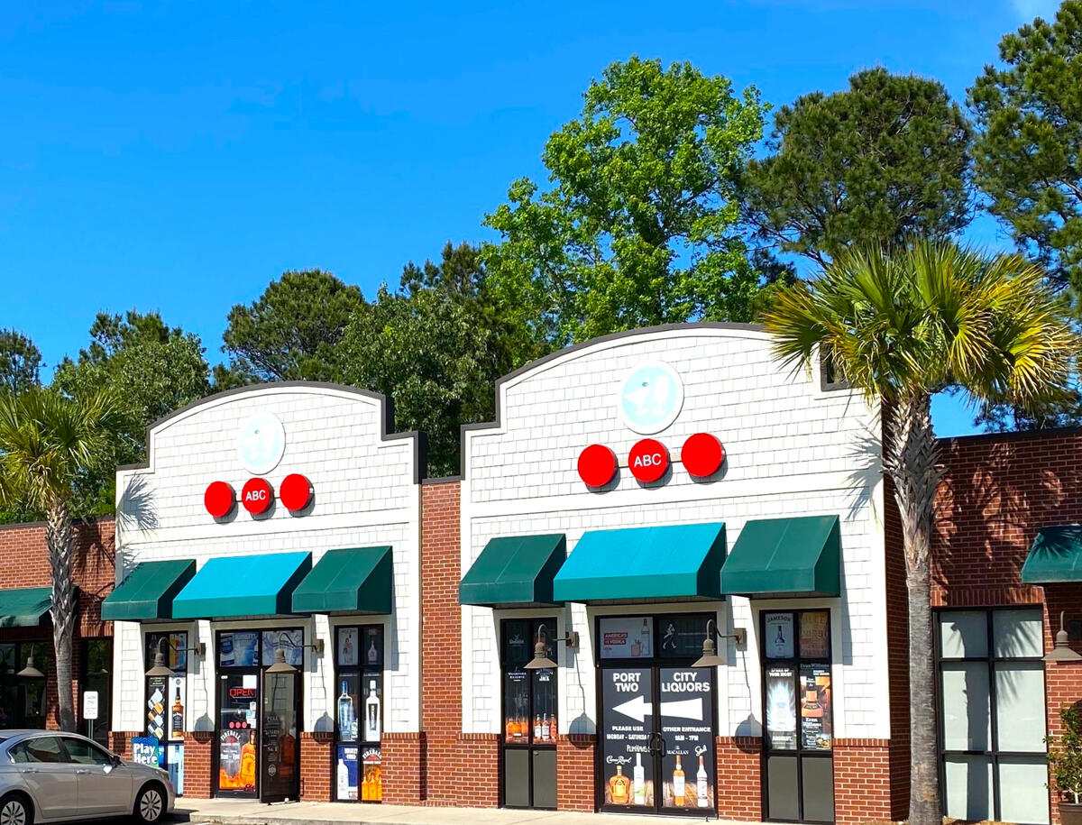 Port City Two Liquors and Wine Storefront – West Ashley, SC Exterior view of Port City Two Liquors and Wine in a modern shopping plaza in West Ashley. The storefront features large glass windows displaying spirit advertisements, a clean tan facade with stone-base pillars, and three distinctive red circular signs ab